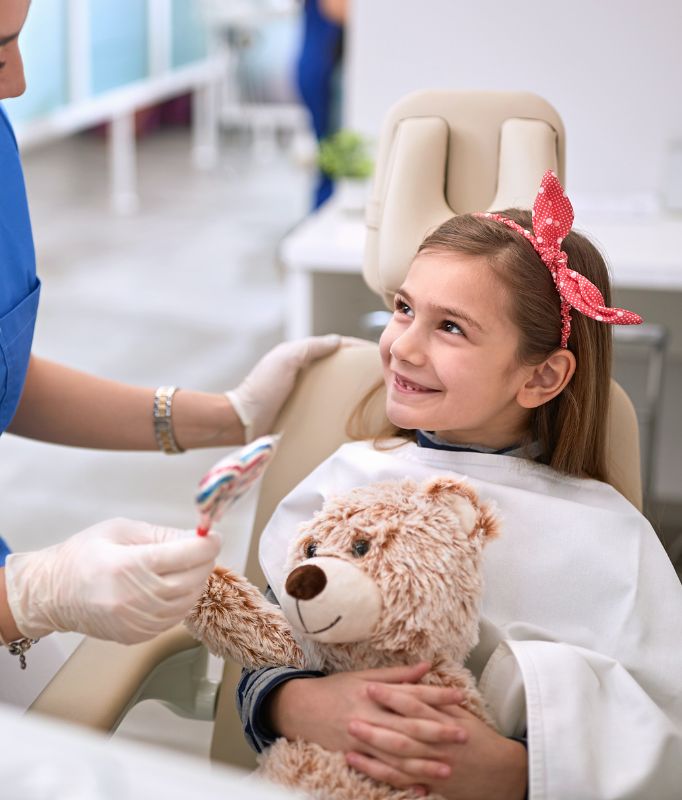 Children's dentistry Kilburn - Young girl sitting in a dentist chair. She is smiling and holding a teddy bear.