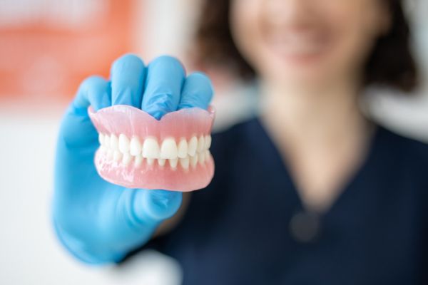 Dental professional, wearing blue surgical gloves, holding a pair of dentures towards the camera.